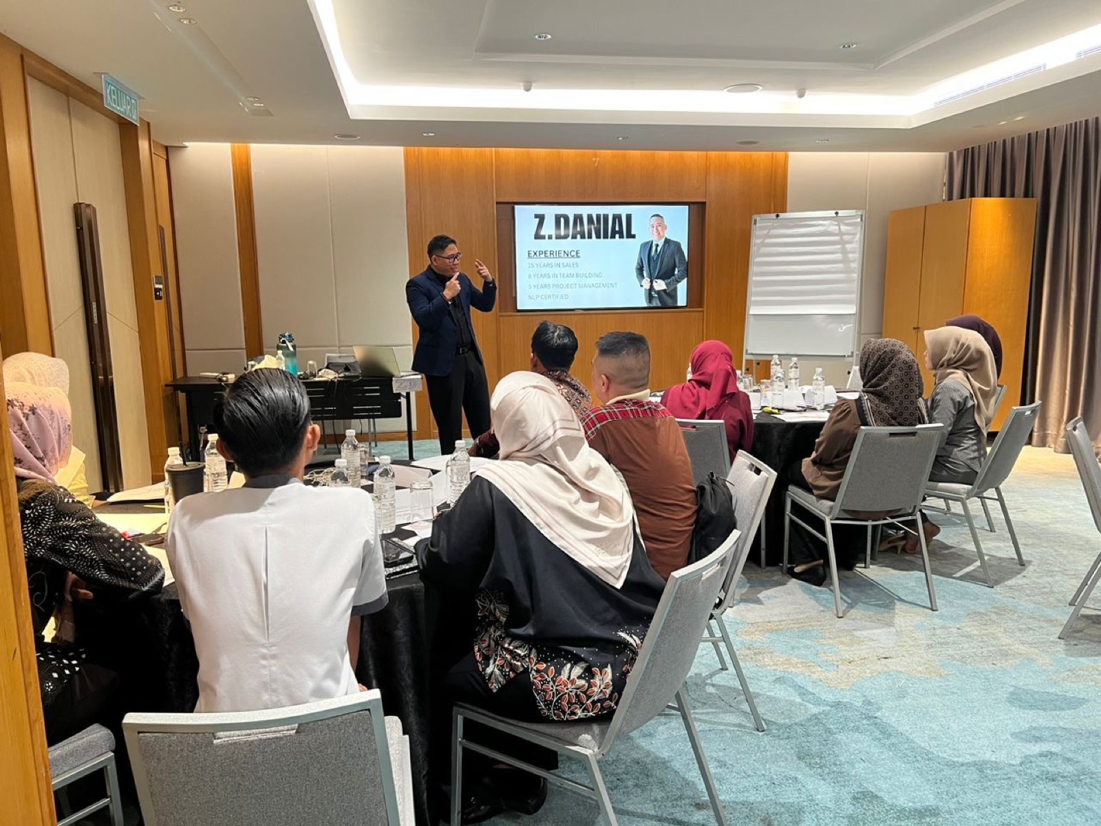 A trainer presenting to participants seated at tables during a public course in a hotel function room